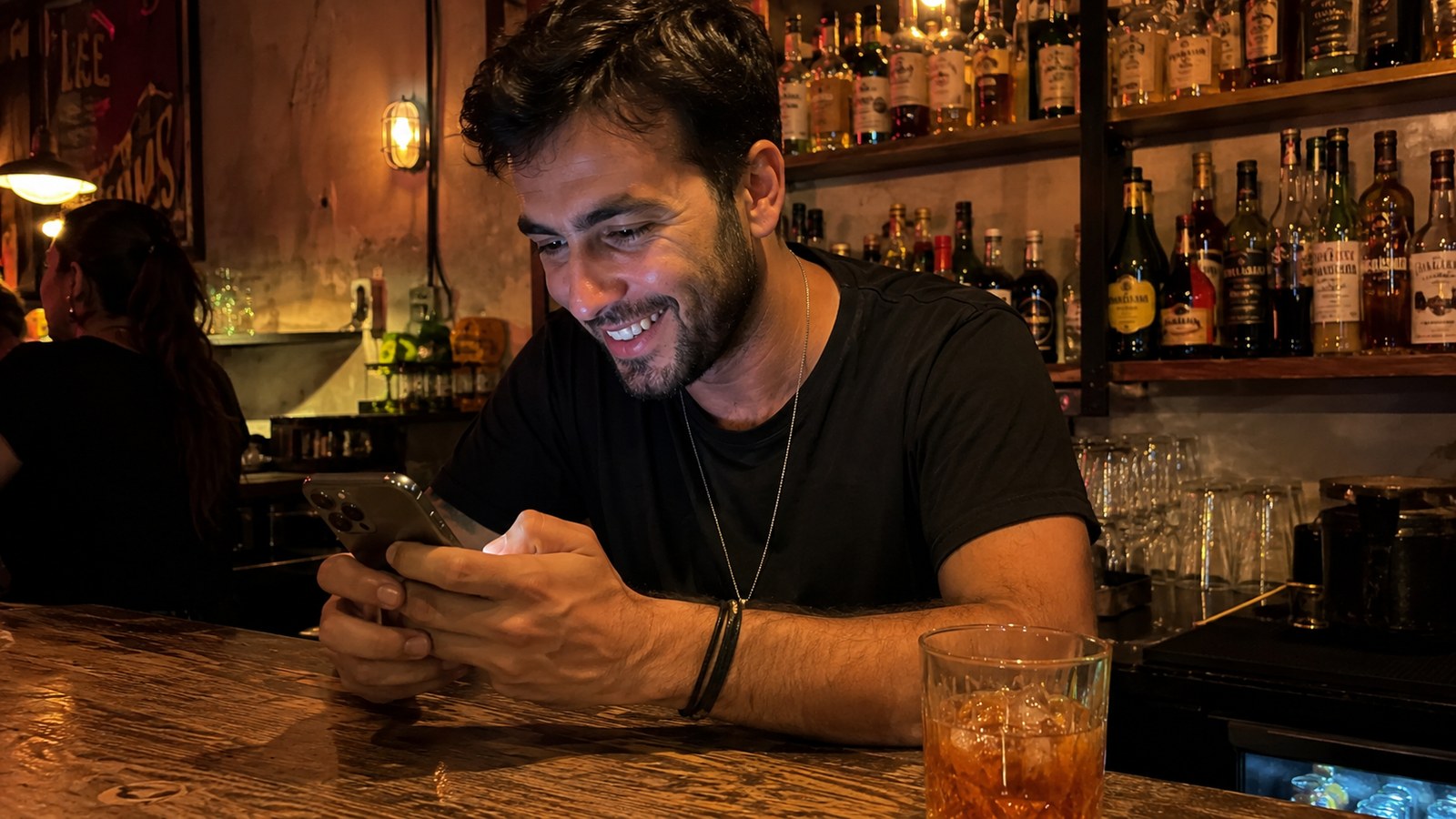 A man at a bar counter typing a message on his phone with a playful grin