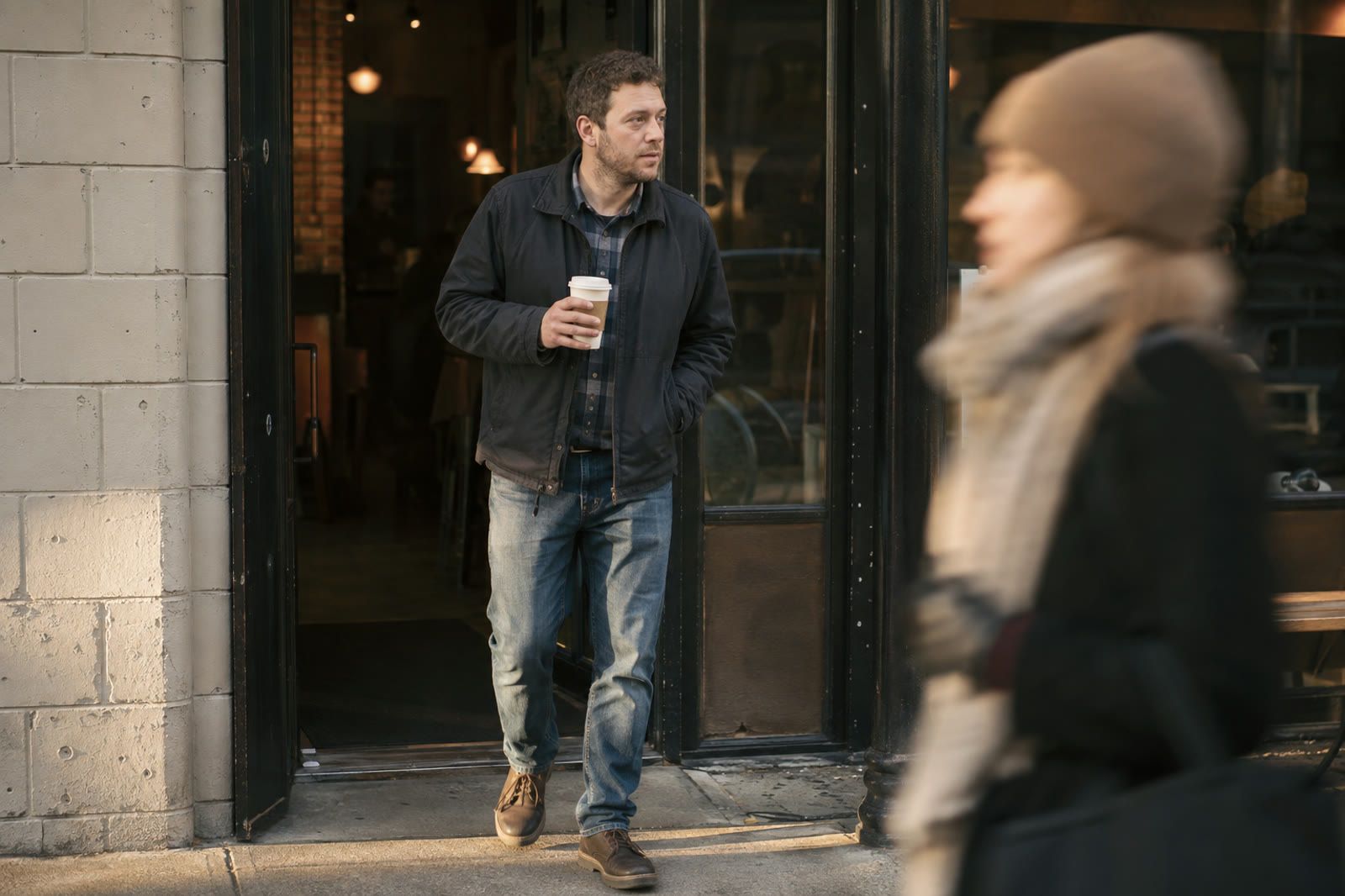 An ordinary man stepping out of a coffee shop with a paper cup, shot like a press photograph while nothing is happening