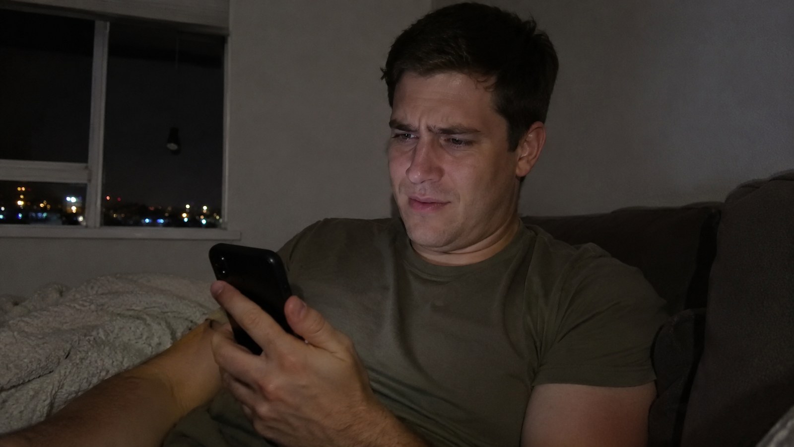 A man on his couch at night staring at his phone, confused and disappointed