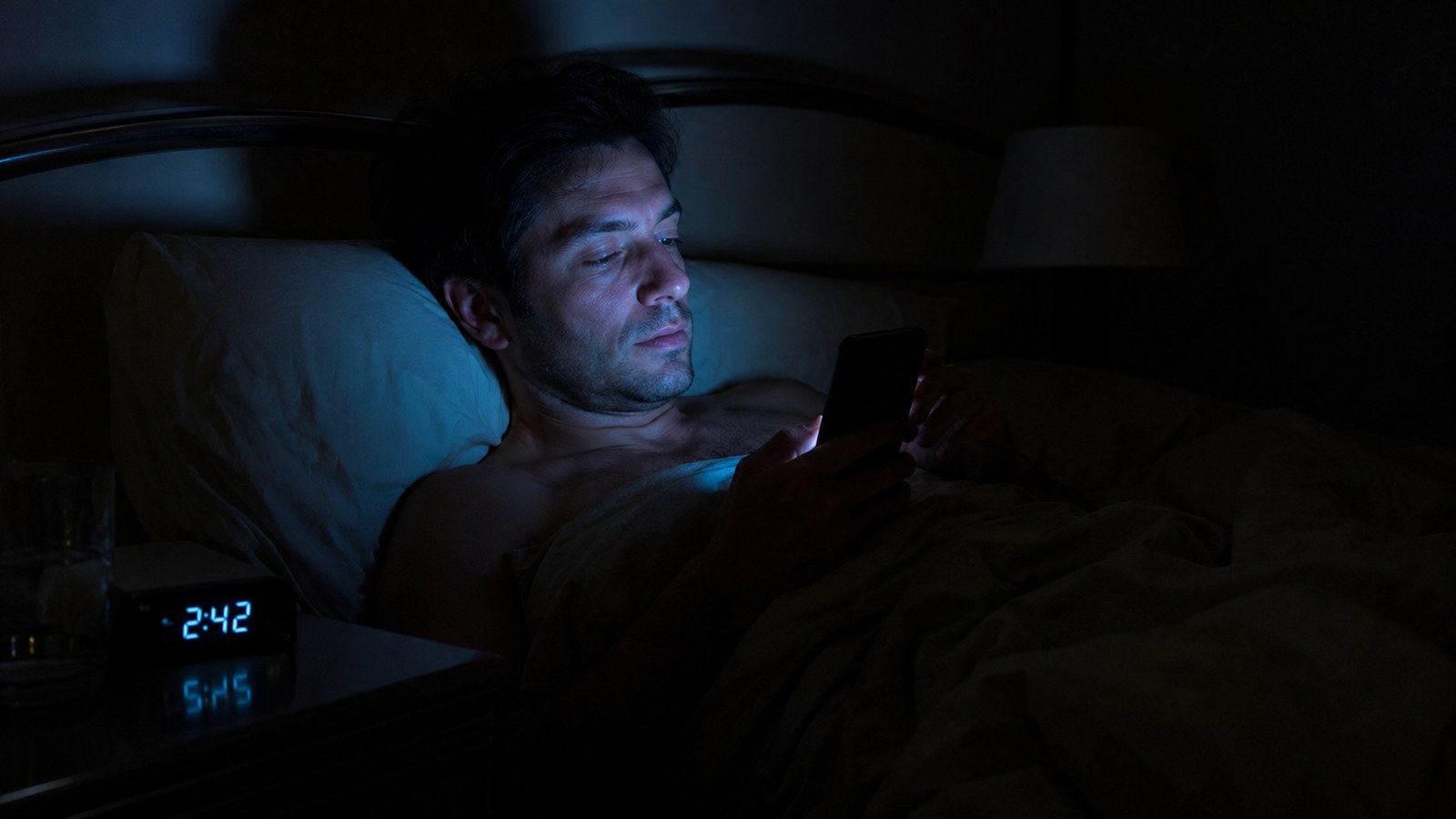 A man in bed at night, face lit by his phone screen, scrolling
