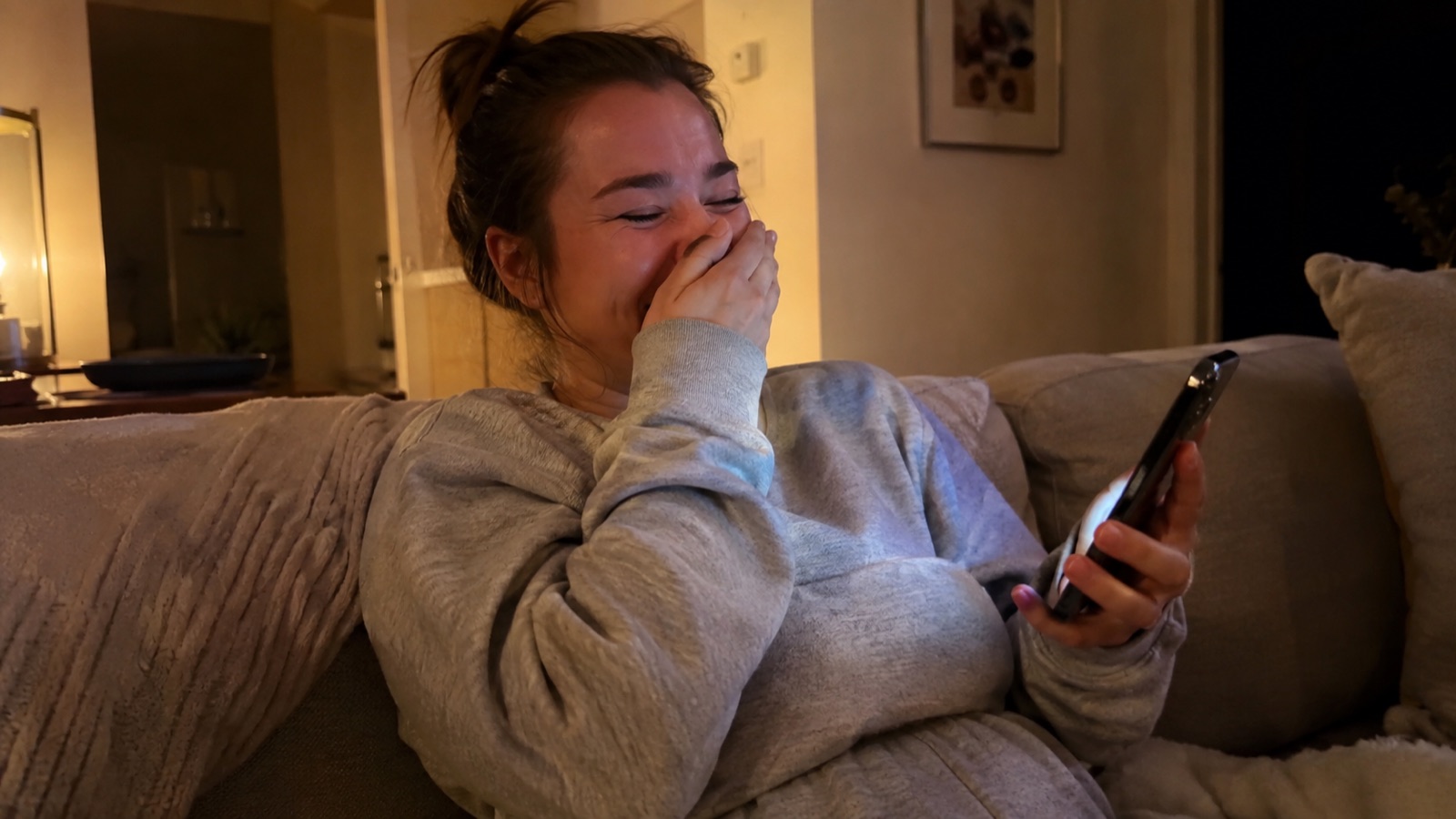 A woman laughing out loud at her phone on a couch, lit by warm evening light