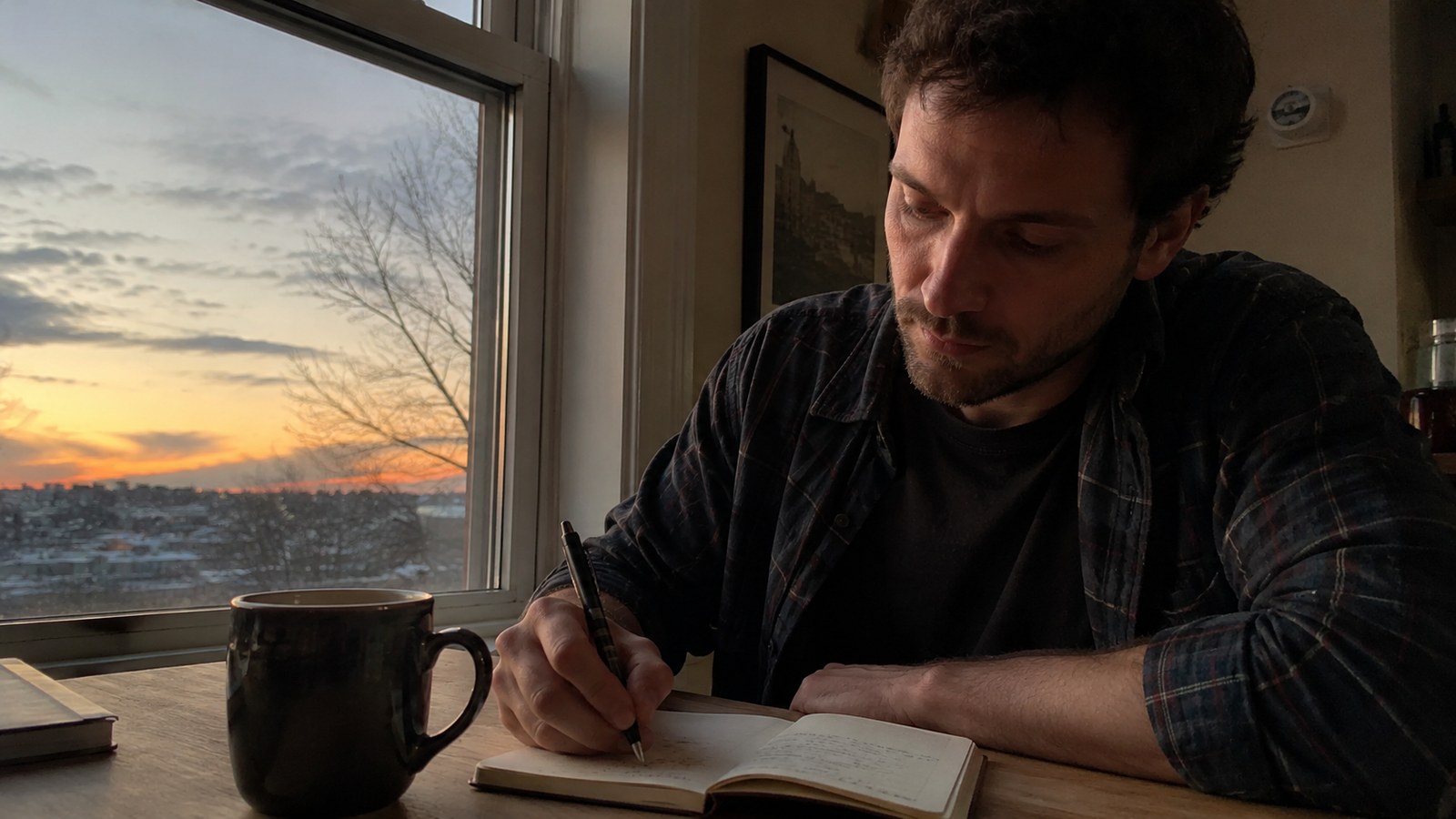 A man writing in a notebook by a window at dusk, calm and thoughtful