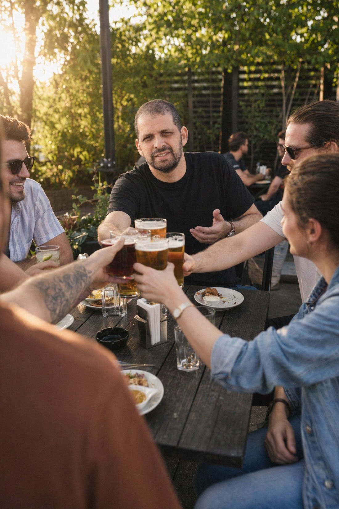 The in-between format: golden hour, cold glasses, real table. Still beats a paper cup on a bench. Man raising glasses with friends at a golden-hour beer garden
