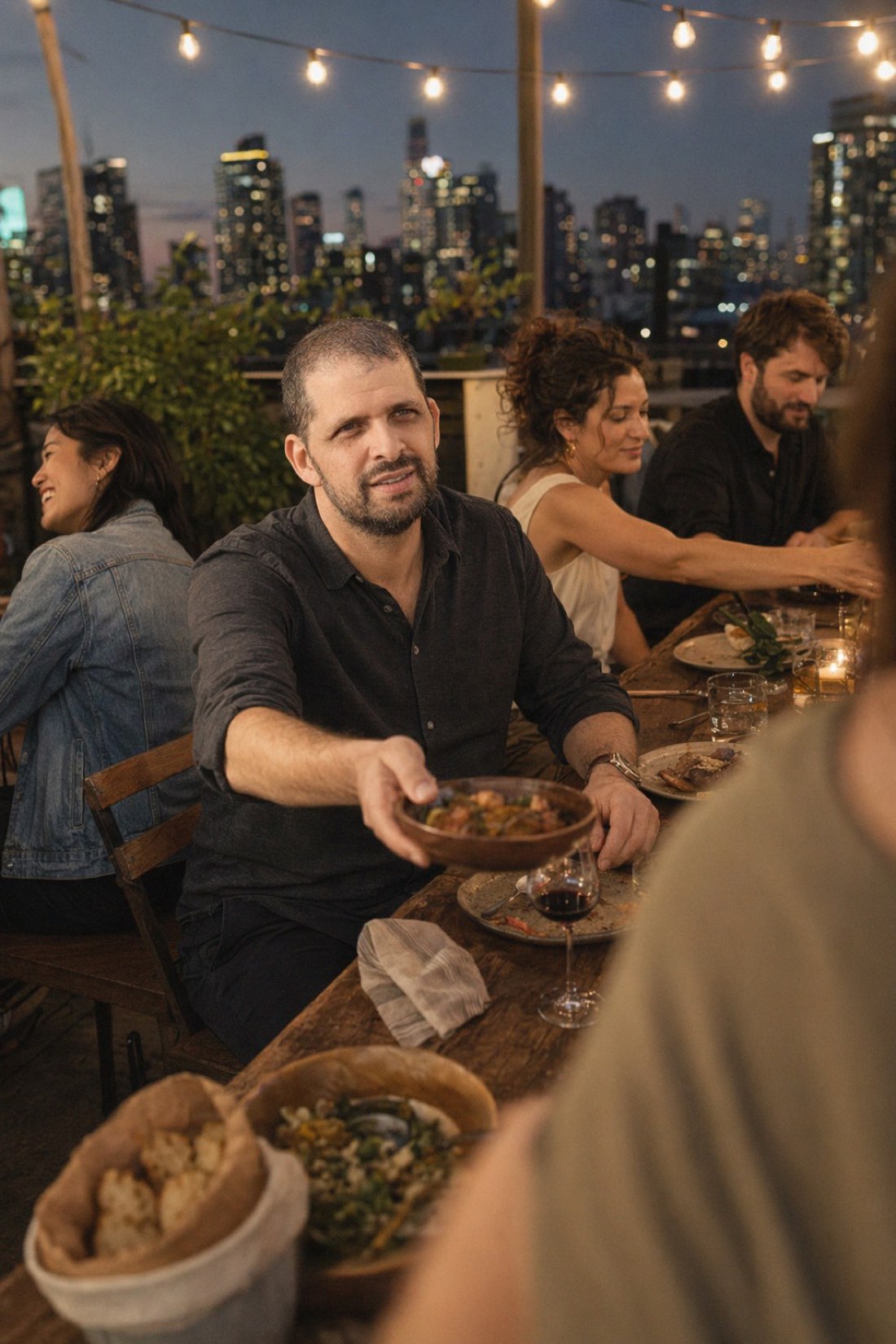 The table does half the work: light, food, time, and something to pass. Chemistry loves logistics. Man passing a dish at a rooftop dinner table under string lights
