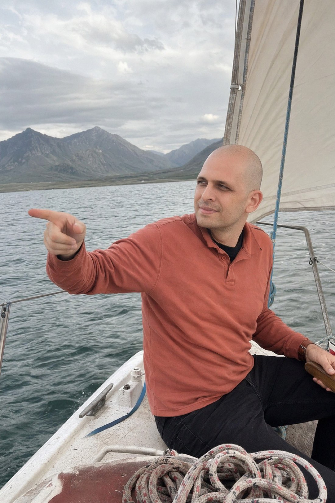Man pointing ahead from a sailboat below mountains, mid-adventure and clearly not waiting by the phone