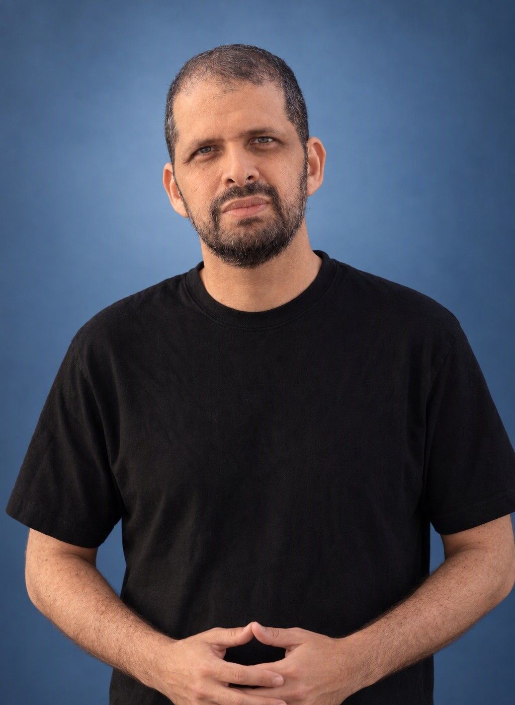 A clear, natural photo where your face reads plainly. That is what a headshot needs to do. AI headshot of a man standing with his hands together against a blue studio backdrop, his real face kept